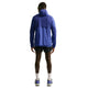 En person står med ryggen til i en Nike Stride Plus løbejakke, Herre, Deep Royal/Lapis/Reflective Silver, sammen med sorte shorts, hvide sokker og løbesko. Baggrunden er almindelig hvid.