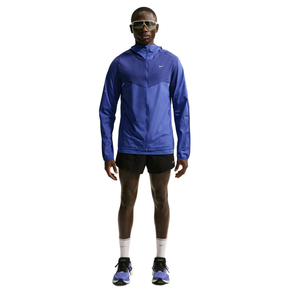 En person står med ansigtet fremad i en Nike Stride Plus løbejakke, Herre (Deep Royal/Lapis/Reflective Silver) med hætte, parret med sorte shorts, hvide sokker, løbesko og solbriller på en almindelig hvid baggrund.