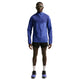 En person står med ansigtet fremad i en Nike Stride Plus løbejakke, Herre (Deep Royal/Lapis/Reflective Silver) med hætte, parret med sorte shorts, hvide sokker, løbesko og solbriller på en almindelig hvid baggrund.