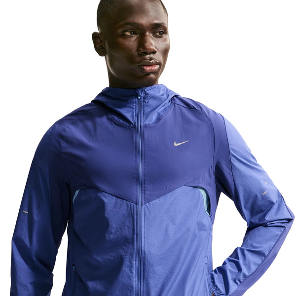 En mand iført en blå Nike Stride Plus løbejakke, Herre, Deep Royal/Lapis/Reflective Silver med hætte står med en hånd på hoften og kigger til siden mod en hvid baggrund.