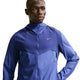 En mand iført en blå Nike Stride Plus løbejakke, Herre, Deep Royal/Lapis/Reflective Silver med hætte står med en hånd på hoften og kigger til siden mod en hvid baggrund.