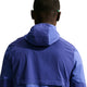 En person ses bagfra iført en Nike Stride Plus Running Jacket, Herre, i Deep Royal/Lapis/Reflective Silver med hætten oppe. Den glatte, vandafvisende jakke skiller sig ud mod en almindelig hvid baggrund.