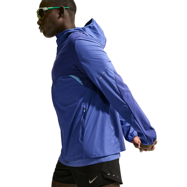 En person iført en blå Nike Stride Plus løbejakke, Herre, Deep Royal/Lapis/Reflective Silver, sorte Nike-shorts og solbriller strækker sig mod en hvid baggrund. Denne vandafvisende løbejakke herre er ideel til enhver træning.