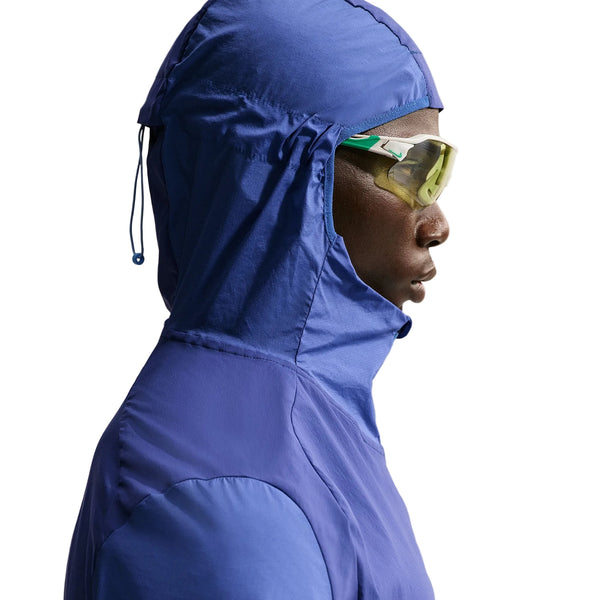 En mand i profil bærer en blå Nike Stride Plus Running Jacket, Herre, Deep Royal/Lapis/Reflective Silver, vandafvisende løbejakke herre, og gule sportssolbriller på en almindelig hvid baggrund.