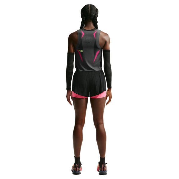 En person med flettet hår står med ryggen til og har en Nike AeroSwift Cropped Running Tank Top til kvinder i farvekombinationen Iron Grey/Volt/White på – en højtydende løbetop til kvinder – kombineret med sorte shorts med lyserøde kanter, sorte armbeskyttere og sorte sneakers med lyserøde detaljer, alt sammen på en hvid baggrund. Outfittet er udstyret med Nikes avancerede Dri-FIT ADV-teknologi, der holder dig kølig og komfortabel ved hvert skridt.
