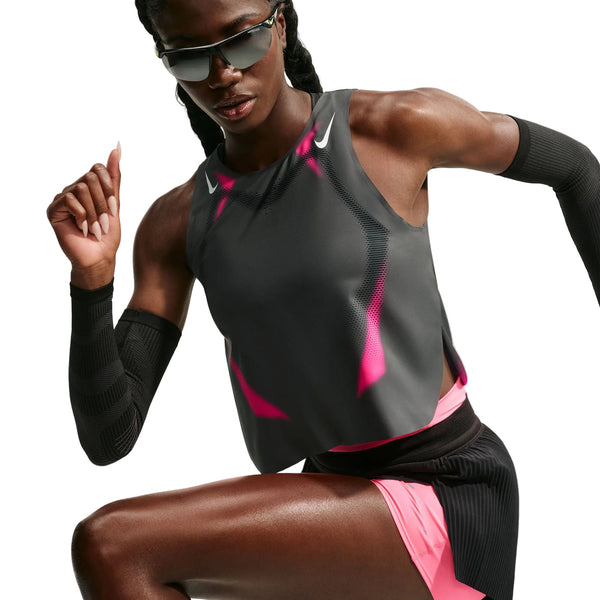 En person iført solbriller, en ærmeløs Nike AeroSwift Cropped løbetop til kvinder i farvekombinationen Iron Grey/Volt/White, matchende shorts og sorte armvarmere poserer i en løbestilling foran en hvid baggrund. Denne stilfulde top er udstyret med Nikes avancerede Dri-FIT ADV-teknologi, der sikrer optimal komfort under træningen.