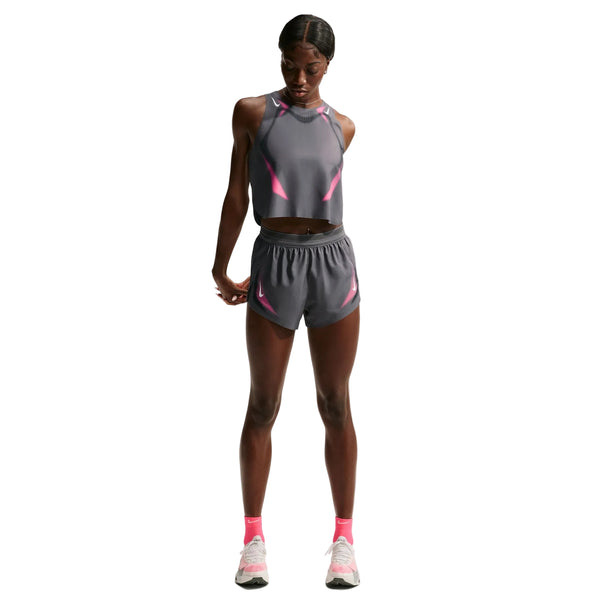 En kvinde står og kigger ned, iført en grå, ærmeløs crop-top og matchende Nike AeroSwift 3In Shorts Gx til kvinder i farven Iron Grey/White med lyserøde detaljer. Hun fuldender looket med lyserøde sokker og hvide sportssko, der fremstår tydeligt mod en ensfarvet hvid baggrund. Disse Nike-løbeshorts er designet med Dri-FIT ADV-teknologi for at give øget komfort under bevægelse.