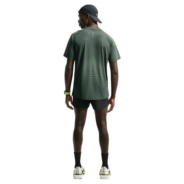 En mand står med ryggen til og er iført en mørk kasket, sorte shorts, sorte sokker, hvide løbesko og et ur på venstre håndled. Han har en Nike Stride Aero-FIT kortærmet løbetrøje til mænd i farvekombinationen sort/lysegrøn Spark/reflekterende sølv på – et design, der sikrer optimal ventilation. Den ensfarvede hvide baggrund fremhæver det sporty Nike-look.