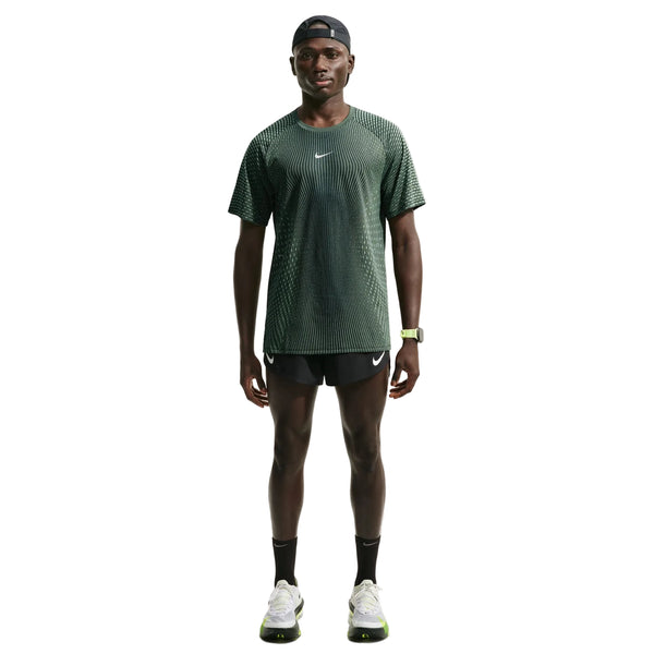 En mand står med ansigtet vendt fremad iført en Nike Stride Aero-FIT kortærmet løbetrøje til mænd i farverne sort/lysegrøn Spark/reflekterende sølv, kombineret med sorte Nike-shorts, sorte sokker, løbesko, en sort kasket og et smartwatch. Baggrunden er ensfarvet hvid.