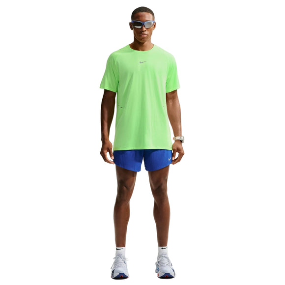 En person står med ansigtet vendt fremad og har en neongrøn Nike Stride Aero-FIT kortærmet løbetrøje til mænd i farverne Volt/Light Silver/Reflective Silver på, der er designet til maksimal ventilation. Outfitet er kombineret med blå shorts, hvide sneakers, et ur og solbriller. Baggrunden er ensfarvet hvid.