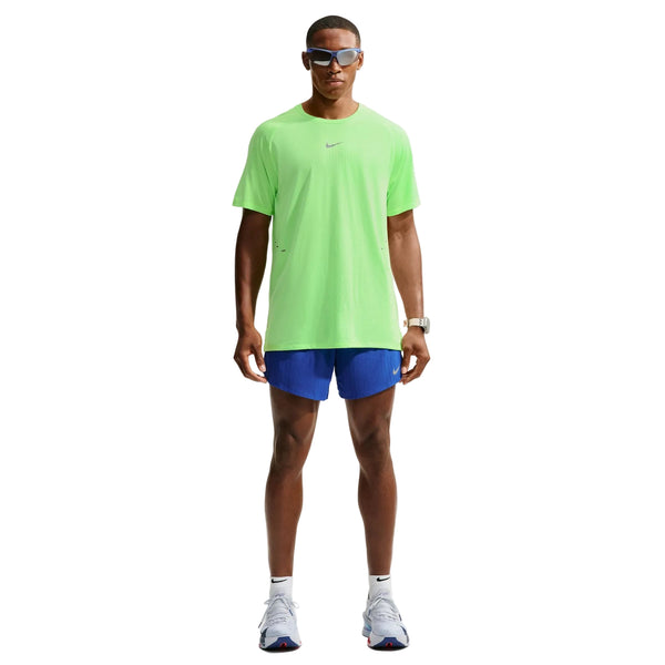 En person står med ansigtet vendt fremad og har en neongrøn Nike Stride Aero-FIT kortærmet løbetrøje til mænd i farverne Volt/Light Silver/Reflective Silver på, der er designet til maksimal ventilation. Outfitet er kombineret med blå shorts, hvide sneakers, et ur og solbriller. Baggrunden er ensfarvet hvid.