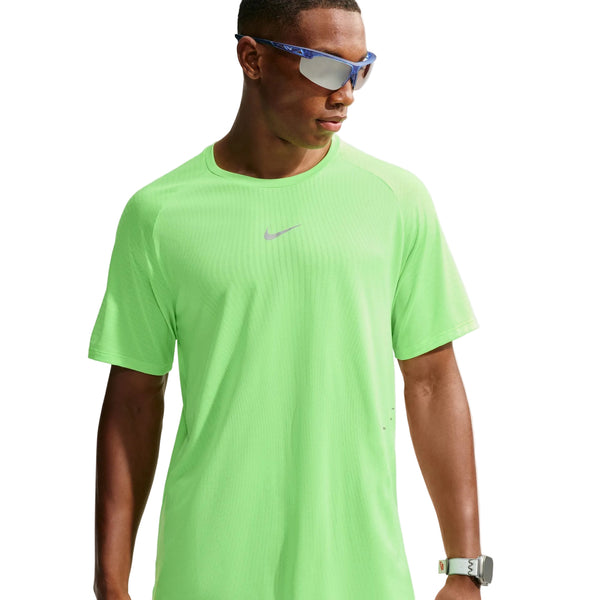 En person iført reflekterende solbriller, en neongrøn Nike Stride Aero-FIT kortærmet løbetrøje til mænd i farverne Volt/Light Silver/Reflective Silver – kendt for sin optimale ventilation – samt et smartur står foran en hvid baggrund og kigger til siden.