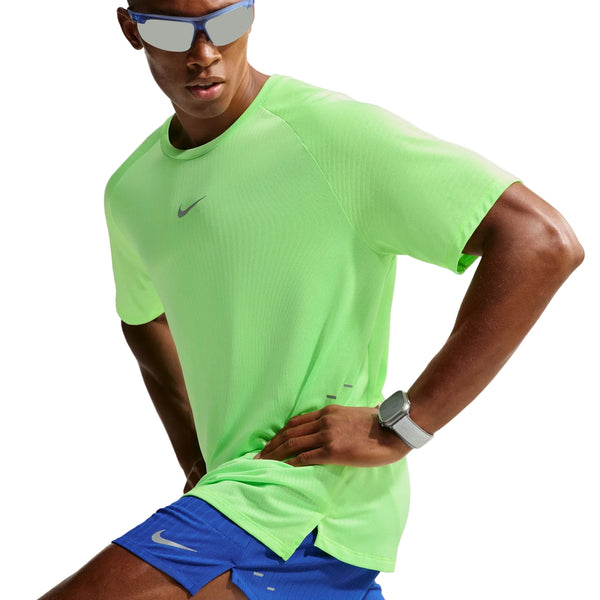 En mand iført en neongrøn (Volt) Nike Stride Aero-FIT kortærmet løbetrøje (Herre, Volt/Light Silver/Reflective Silver), blå Nike-shorts, solbriller og et ur poserer med hænderne på hofterne foran en hvid baggrund. Trøjen er designet til maksimal ventilation og optimal løbekomfort.