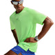 En mand iført en neongrøn (Volt) Nike Stride Aero-FIT kortærmet løbetrøje (Herre, Volt/Light Silver/Reflective Silver), blå Nike-shorts, solbriller og et ur poserer med hænderne på hofterne foran en hvid baggrund. Trøjen er designet til maksimal ventilation og optimal løbekomfort.
