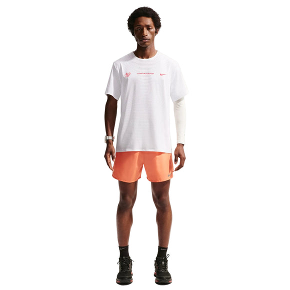 En mand ser frem for sig iført en Summit White Nike Nk Dfadv Aeroswift Gfx Ss T-shirt, orange sportsshorts, sorte sneakers, et hvidt kompressionsærme på venstre arm og et ur på højre håndled mod en almindelig hvid baggrund.