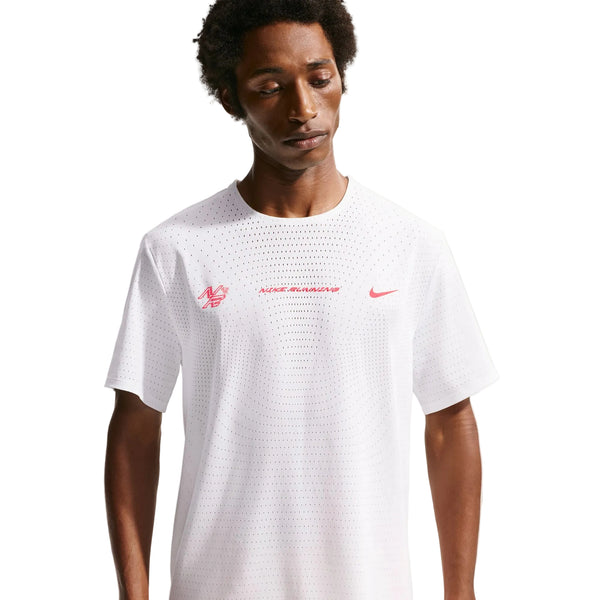 En mand iført Nike Nk Dfadv Aeroswift Gfx Ss i Summit White med Dri-FIT ADV-teknologi og rød brystgrafik står på en almindelig baggrund og kigger ned med et neutralt udtryk.