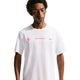 En mand iført Nike Nk Dfadv Aeroswift Gfx Ss i Summit White med Dri-FIT ADV-teknologi og rød brystgrafik står på en almindelig baggrund og kigger ned med et neutralt udtryk.