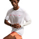 En mand bærer Nike Nk Dfadv Aeroswift Gfx Ss løbetrøje herre i Summit White med pink tekst og logo, orange shorts, et hvidt Nike-armstykke og et smartwatch, mens han står med foldede hænder.