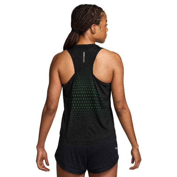 En kvinde med flettet hår ses bagfra, iført Nike Swift Aero-FIT løbetop til kvinder i farvekombinationen sort/lysegrøn Spark/reflekterende sølv samt matchende sorte shorts. Den ærmeløse sportstop er designet til maksimal ventilation og er prydet med små grønne prikker samt teksten »AEROREADY« trykt tæt på kraven. Hun står foran en ensfarvet hvid baggrund.