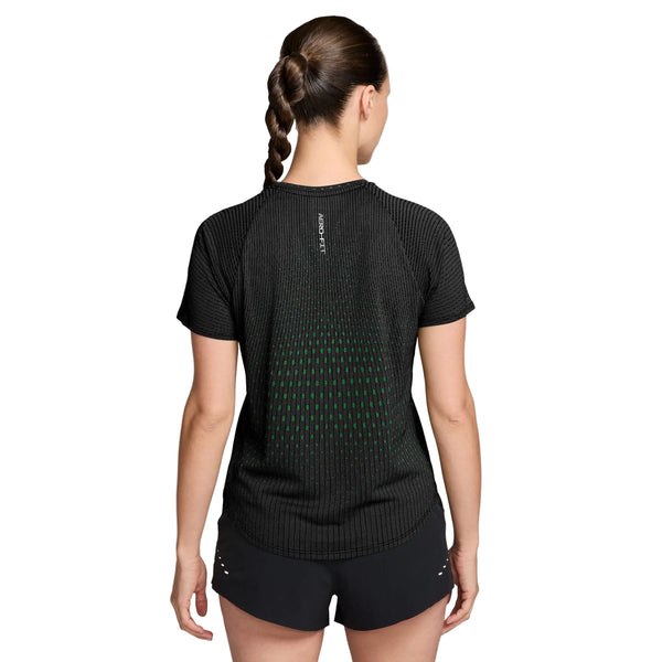 En kvinde med en fletning ses set bagfra iført Nike Swift Aero-FIT kortærmet løbetrøje i farvekombinationen sort/lysegrøn spark/reflekterende sølv, kombineret med sorte shorts. Toppen har et diskret grønt mønster, et lille logo nær halsudskæringen og fremviser Nikes karakteristiske Aero-FIT-design, der sikrer forbedret ydeevne.