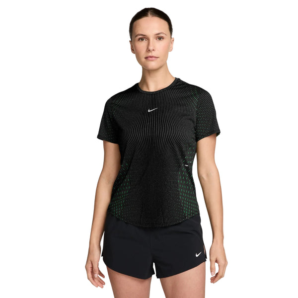 En kvinde står med ansigtet vendt fremad iført Nike Swift Aero-FIT kortærmet løbetrøje til kvinder i farvekombinationen sort/lysegrøn Spark/reflekterende sølv, kombineret med sorte Nike-shorts. Hendes hår er sat op, mens hun poserer foran en ensfarvet hvid baggrund.