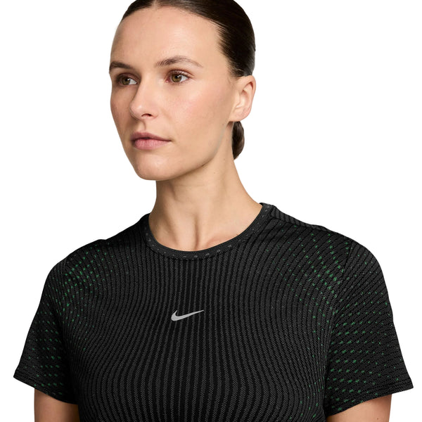 En person med mørkt hår, der er sat op, har en Nike Swift Aero-FIT kortærmet løbetrøje i farverne sort/lysegrøn spark/reflekterende sølv på, med et hvidt Swoosh-logo. Personen kigger let til siden foran en ensfarvet hvid baggrund og udstråler en cool, atletisk kvindelig stil.