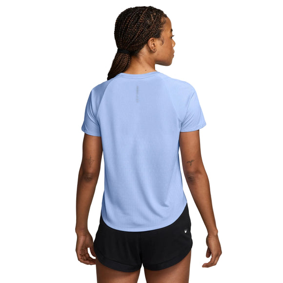 Der ses en person med flettet hår set bagfra, iført en lyseblå Nike Swift Aero-FIT kortærmet løbetrøje til kvinder i farverne Aluminium/Pure Platinum/Reflective Silver samt sorte shorts. Baggrunden er ensfarvet hvid.