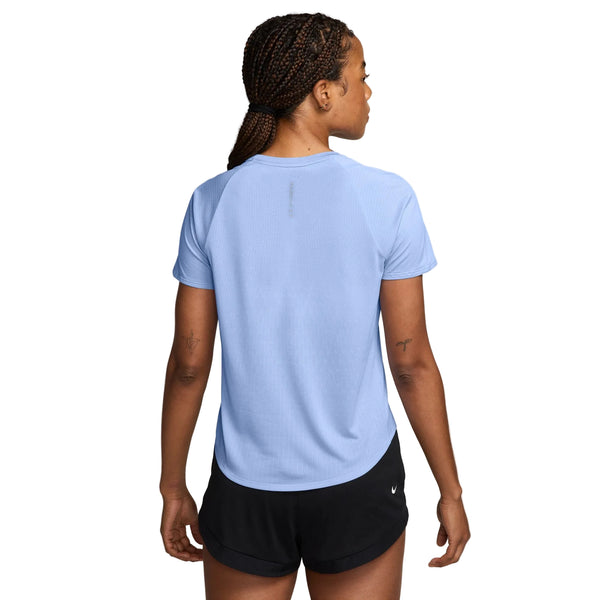 Der ses en person med flettet hår set bagfra, iført en lyseblå Nike Swift Aero-FIT kortærmet løbetrøje til kvinder i farverne Aluminium/Pure Platinum/Reflective Silver samt sorte shorts. Baggrunden er ensfarvet hvid.