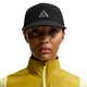 En person iført Nike ACG Fly Cap (Unisex, Black/Black/Black/Black) med en gul vest med lynlås over en lys skjorte, kigger fremad med et neutralt udtryk mod en almindelig baggrund.