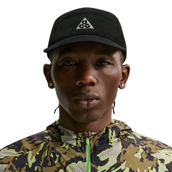 En person iført en sort Nike ACG Fly Cap (Unisex, Black/Black/Black/Black) og en camouflagejakke ser direkte ind i kameraet mod en hvid baggrund.
