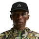 En person iført en sort Nike ACG Fly Cap (Unisex, Black/Black/Black/Black) og en camouflagejakke ser direkte ind i kameraet mod en hvid baggrund.