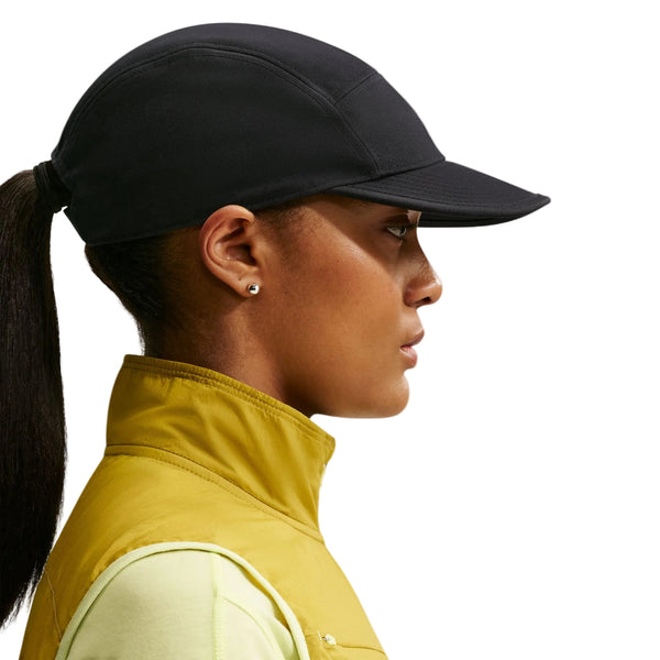 En person med hestehale bærer Nike ACG Fly Cap (Unisex, sort) med ørestikker, en gul vest med høj krave og en lys skjorte, vist i profil mod en hvid baggrund.