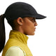En person med hestehale bærer Nike ACG Fly Cap (Unisex, sort) med ørestikker, en gul vest med høj krave og en lys skjorte, vist i profil mod en hvid baggrund.