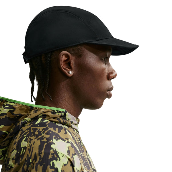 En person i profil mod højre bærer en sort Nike ACG Fly Cap (Unisex, Black/Black/Black/Black), en camouflagejakke med grøn kant, små øreringe og har korte fletninger.