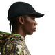 En person i profil mod højre bærer en sort Nike ACG Fly Cap (Unisex, Black/Black/Black/Black), en camouflagejakke med grøn kant, små øreringe og har korte fletninger.