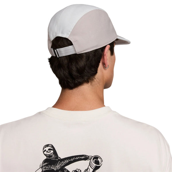 En person med kort mørkt hår og en ørering ses bagfra iført en Nike ACG Fly Cap (Unisex, Photon Dust/College Grey/Photon Dust) og en hvid skjorte med en sort illustration af en figur, der holder en genstand, på ryggen.