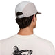 En person med kort mørkt hår og en ørering ses bagfra iført en Nike ACG Fly Cap (Unisex, Photon Dust/College Grey/Photon Dust) og en hvid skjorte med en sort illustration af en figur, der holder en genstand, på ryggen.