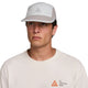 En person bærer en Nike ACG Fly Cap i Photon Dust/College Grey, unisex, sammen med en cremefarvet skjorte med et orange ACG-logo og teksten "All Conditions Gear". Baggrunden er almindelig hvid.