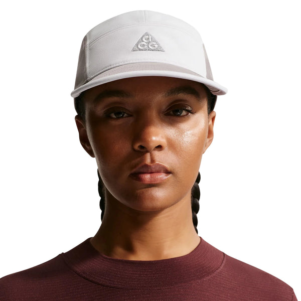 En person med flettet hår bærer en Nike ACG Fly Cap (Unisex, Photon Dust/College Grey) med et trekantet logo og en rødbrun top med rund hals, mens han vender sig mod kameraet med et neutralt udtryk mod en hvid baggrund.