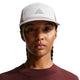 En person med flettet hår bærer en Nike ACG Fly Cap (Unisex, Photon Dust/College Grey) med et trekantet logo og en rødbrun top med rund hals, mens han vender sig mod kameraet med et neutralt udtryk mod en hvid baggrund.