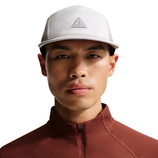 En person kigger fremad mod en hvid baggrund iført en unisex Nike ACG Fly Cap i Photon Dust/College Grey med broderet trekantlogo og vandafvisende finish, sammen med en rustfarvet top med lynlås.