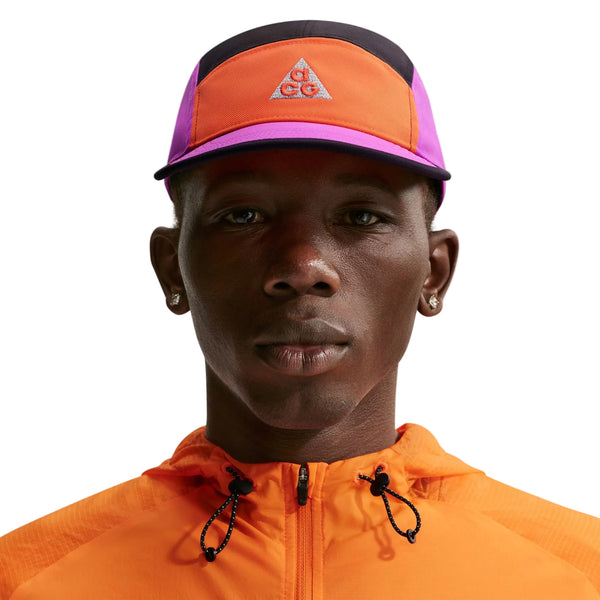 En person bærer en Nike ACG Fly Cap Unisex i Safety Orange/Vivid Purple/Safety Orange med et trekantet logo sammen med en orange jakke med lynlås og snøre, vist på en hvid baggrund.