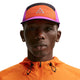 En person bærer en Nike ACG Fly Cap Unisex i Safety Orange/Vivid Purple/Safety Orange med et trekantet logo sammen med en orange jakke med lynlås og snøre, vist på en hvid baggrund.
