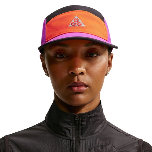En person er iført en sort jakke med lynlås og en Nike ACG Fly Cap Unisex i Safety Orange/Vivid Purple/Safety Orange med ACG-logoet på en hvid baggrund.