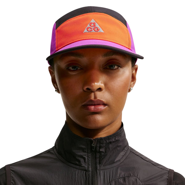 En person er iført en sort jakke med lynlås og en Nike ACG Fly Cap Unisex i Safety Orange/Vivid Purple/Safety Orange med ACG-logoet på en hvid baggrund.