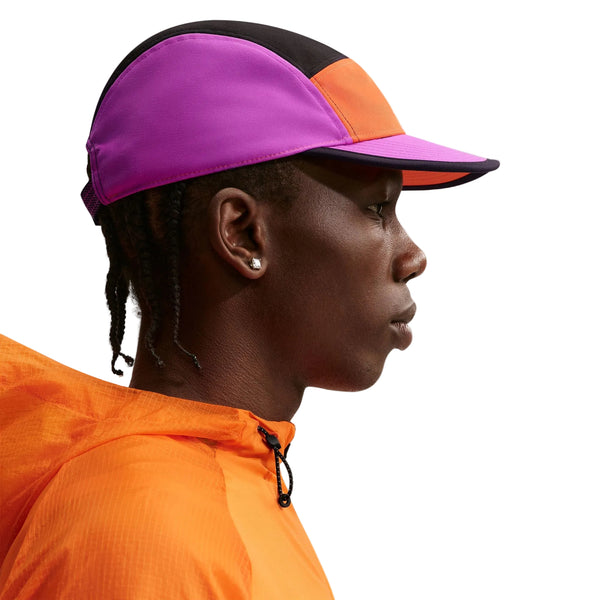 En person i profil bærer Nike ACG Fly Cap Unisex i Safety Orange/Vivid Purple/Safety Orange med en orange jakke. De har kort flettet hår, små ørestikker og er placeret på en almindelig hvid baggrund.