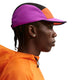 En person i profil bærer Nike ACG Fly Cap Unisex i Safety Orange/Vivid Purple/Safety Orange med en orange jakke. De har kort flettet hår, små ørestikker og er placeret på en almindelig hvid baggrund.