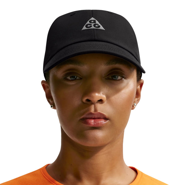 En person, der bærer en Nike ACG Club Unstructured Cap i sort/sort, en orange skjorte og små ørestikker, ser direkte ind i kameraet foran en ensfarvet baggrund.