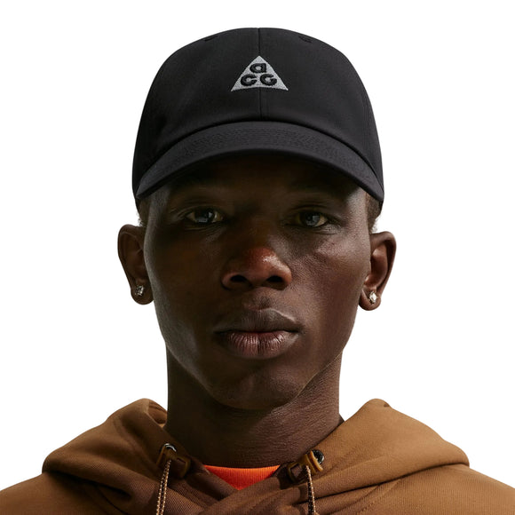 En person iført en sort Nike ACG Club Unstructured Cap med et trekantet logo, en brun hættetrøje og små ringøreringe, der ser fremad foran en ensfarvet hvid baggrund.