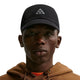 En person iført en sort Nike ACG Club Unstructured Cap med et trekantet logo, en brun hættetrøje og små ringøreringe, der ser fremad foran en ensfarvet hvid baggrund.